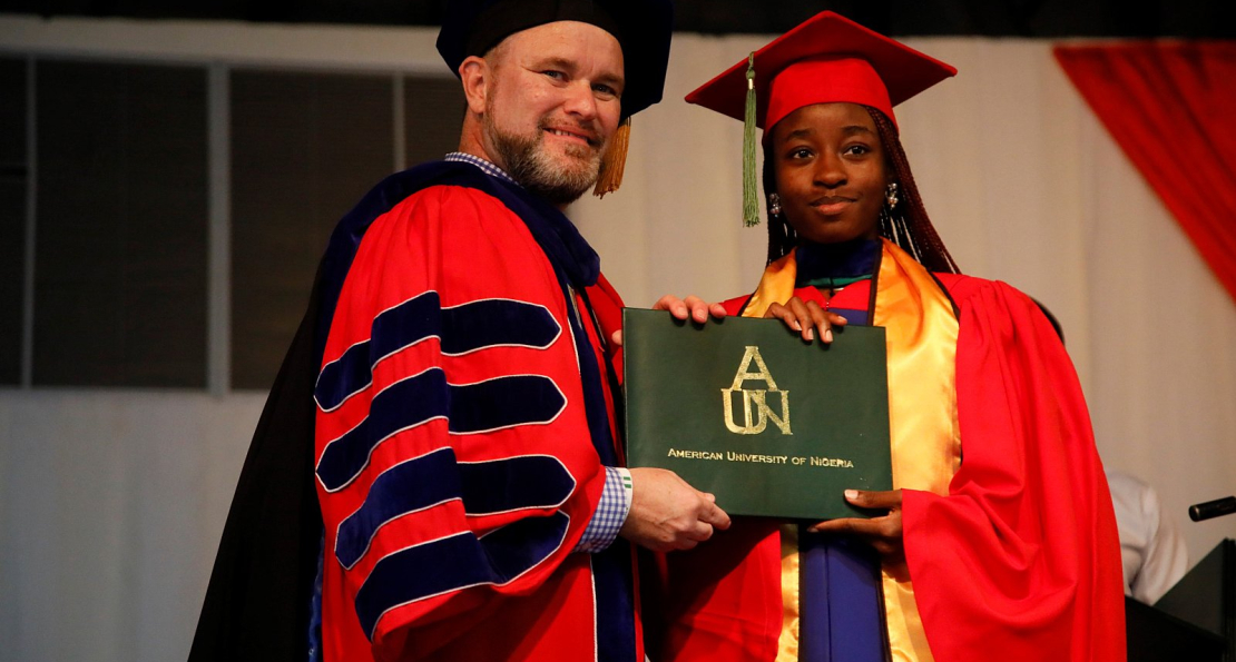 A Nigerian graduate in a red and gold gown receiving a diploma from a university official at the American University of Nigeria graduation ceremony