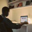 A Nigerian student studying at a desk while using a laptop for freelance work.