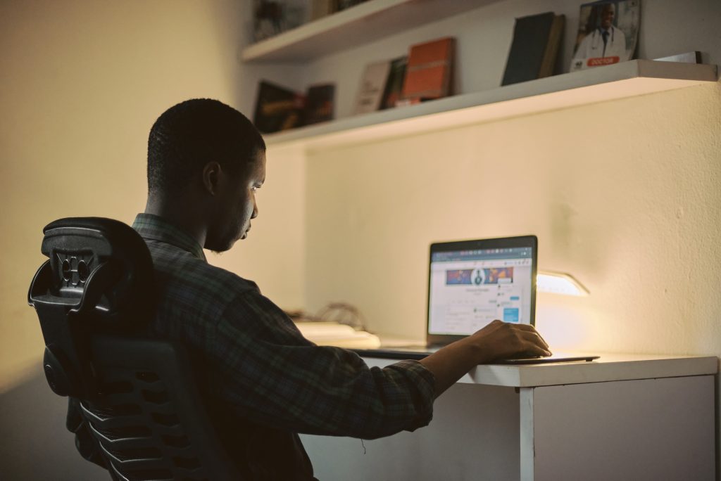 A Nigerian student studying at a desk while using a laptop to balance side hustle with academics without failing.